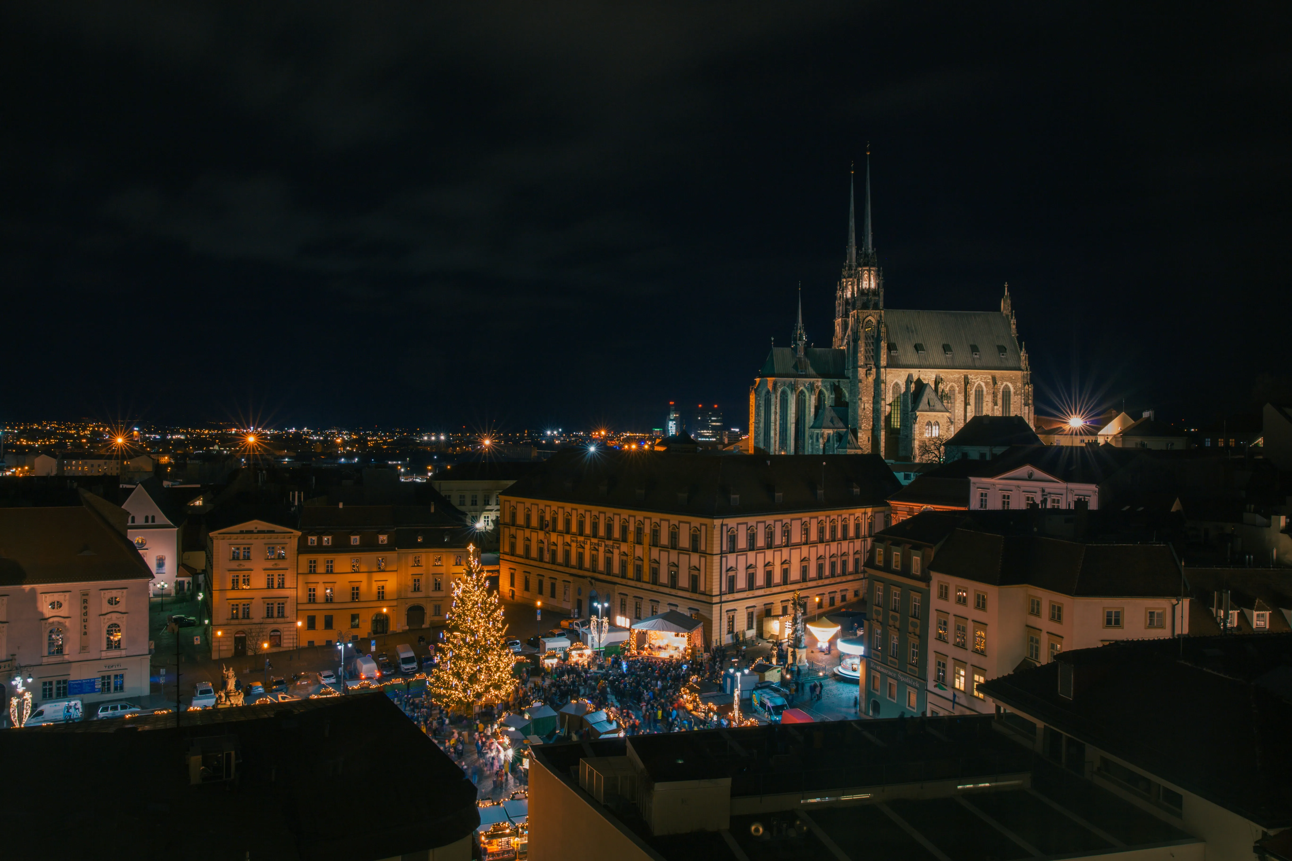 Night view of a crowded, illuminated square in Brno beneath the Cathedral of St Peter and Paul
