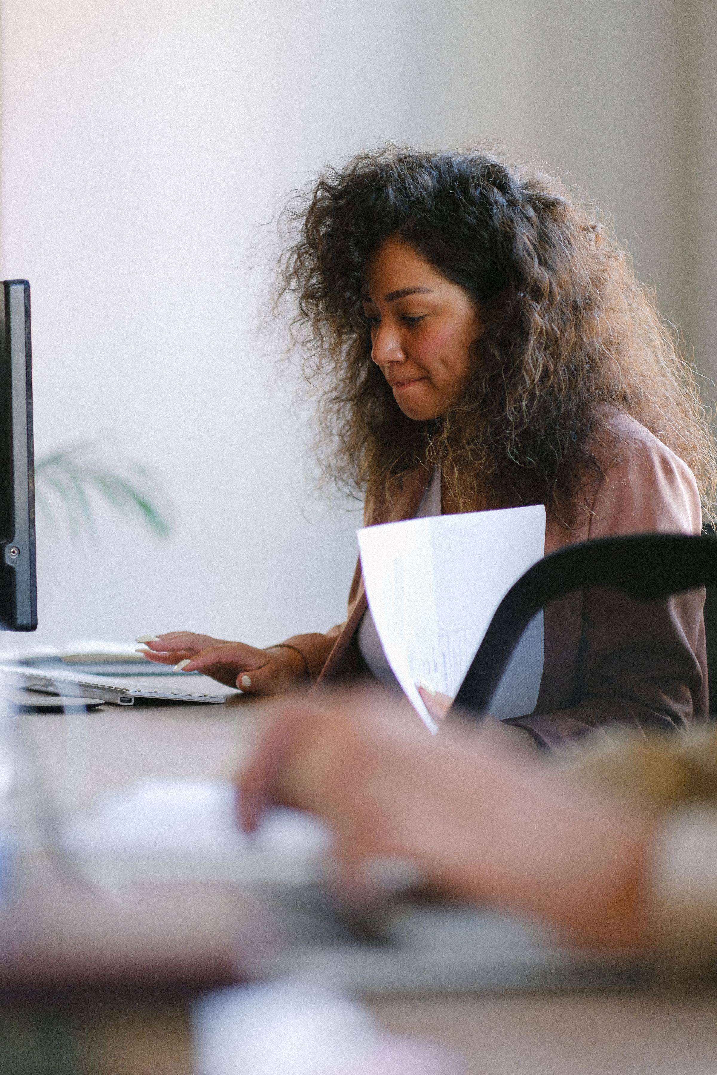 A woman at an office desk holding a printed form and typing at a keyboard in front of a monitor, side view