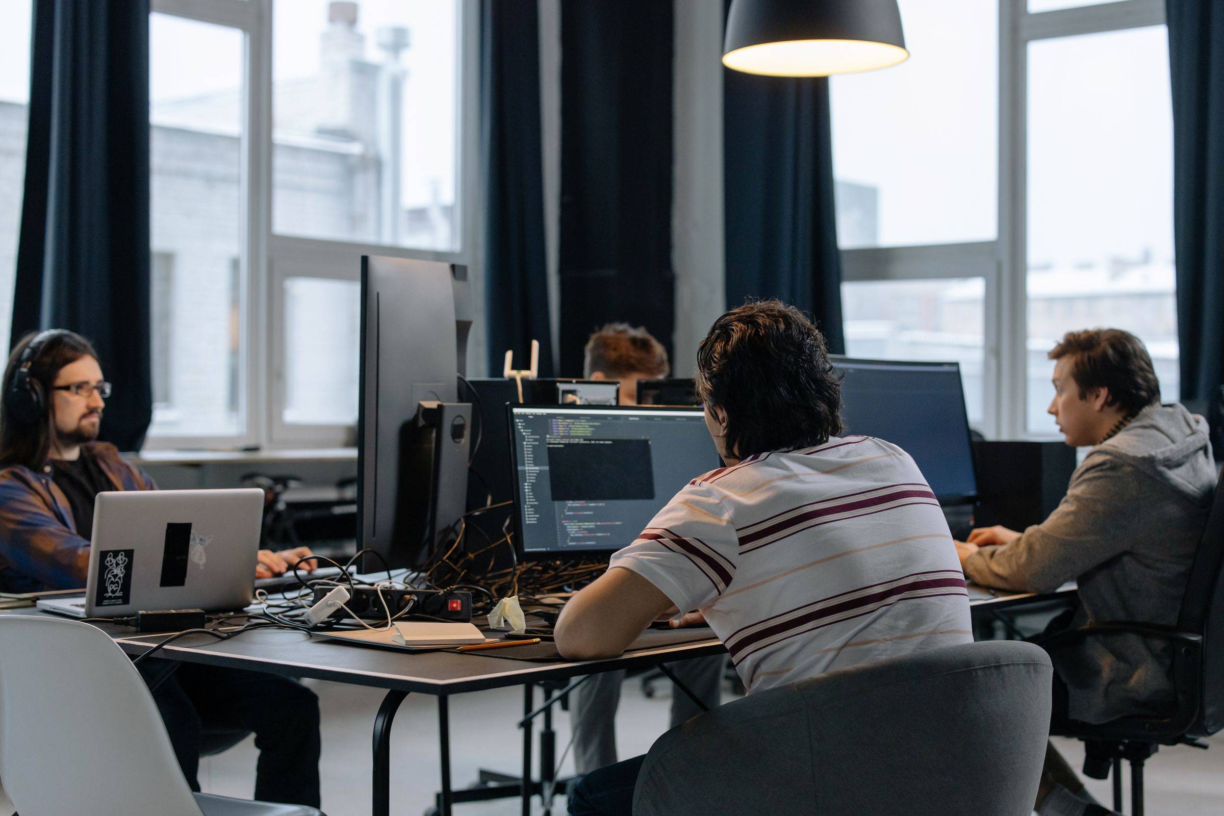 Tři vývojáři u stolů v kanceláři s velkými okny, nejbližší zezadu u monitoru