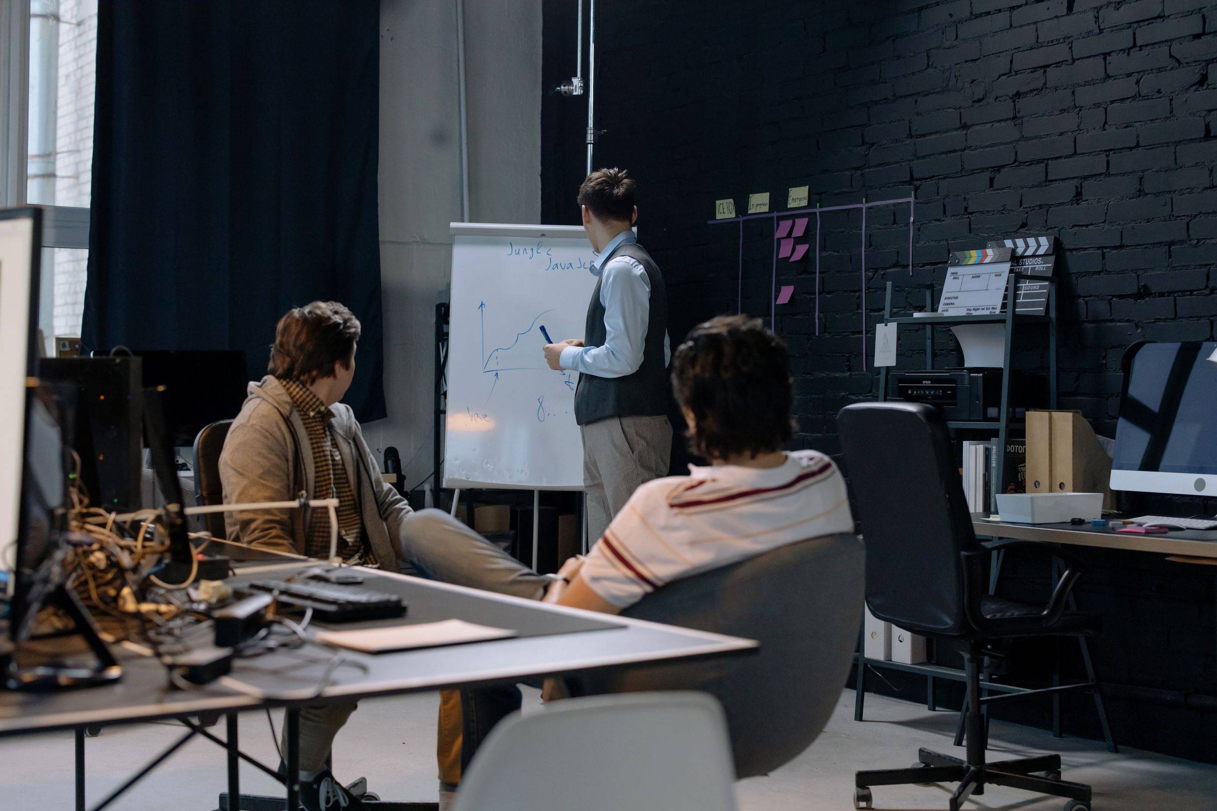 A small team in a dark meeting room following a presenter at a whiteboard
