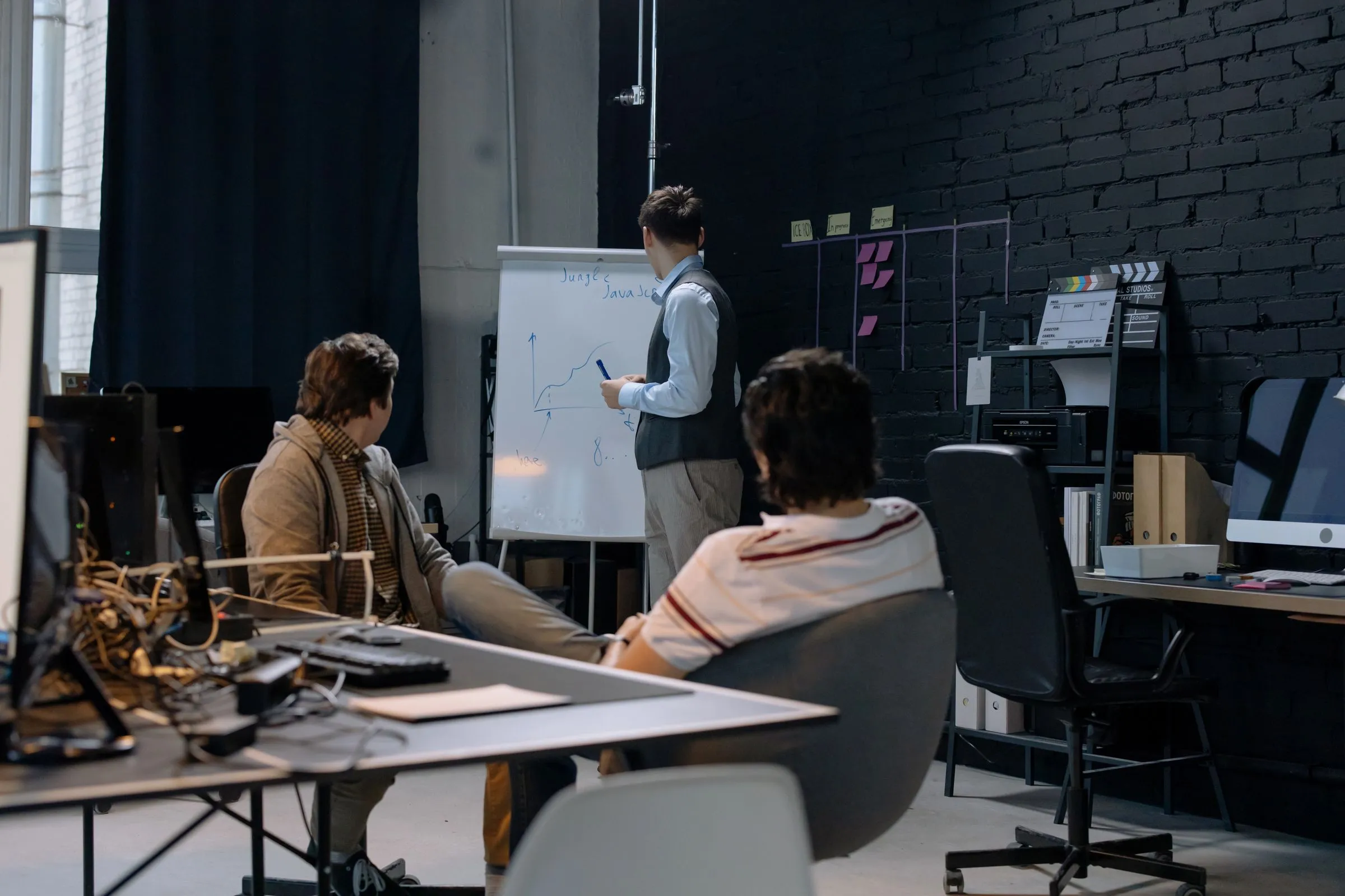 A small team in a dark meeting room following a presenter at a whiteboard
