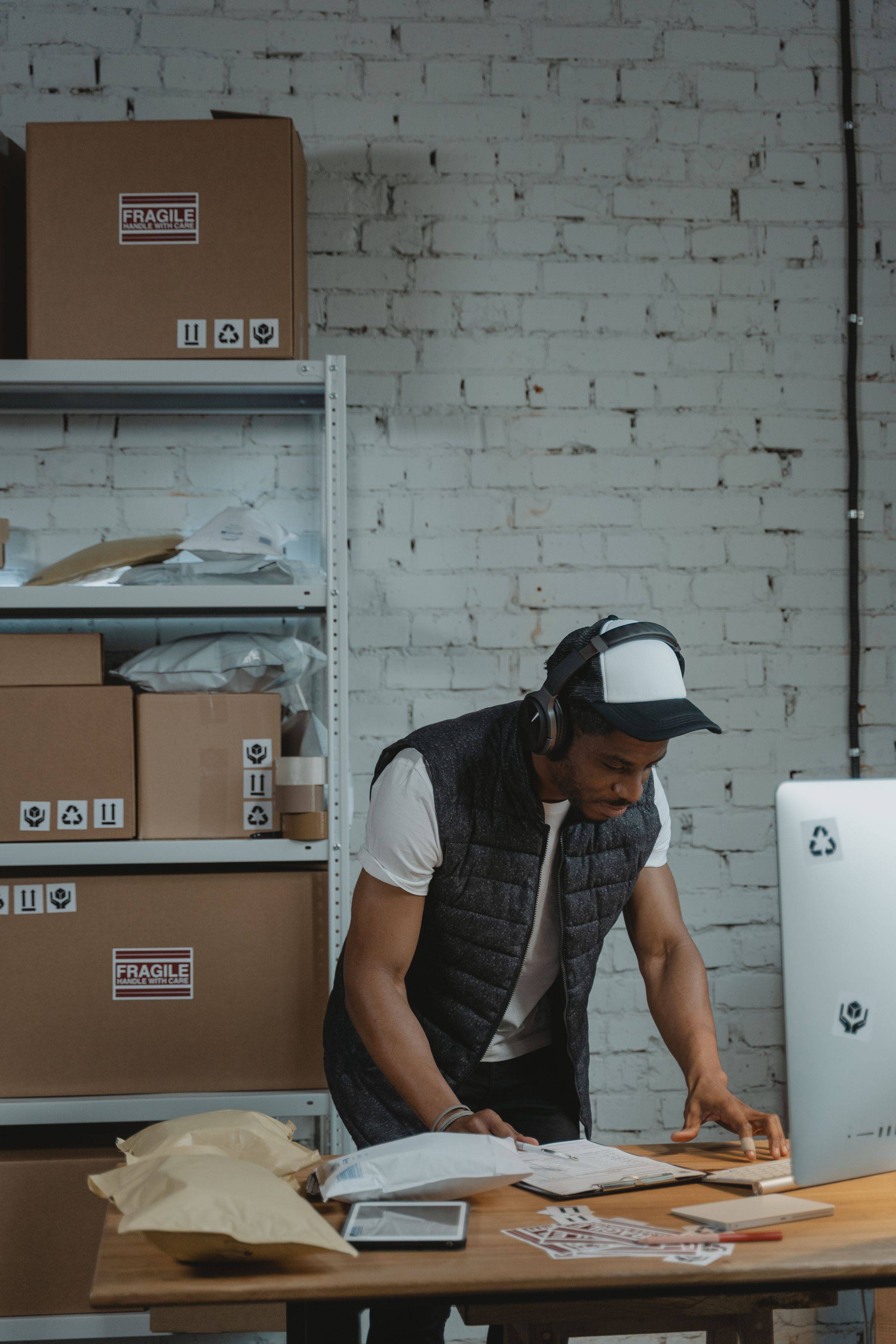 A man in a cap and headphones leans over a desk with paperwork and parcels in a shipping warehouse