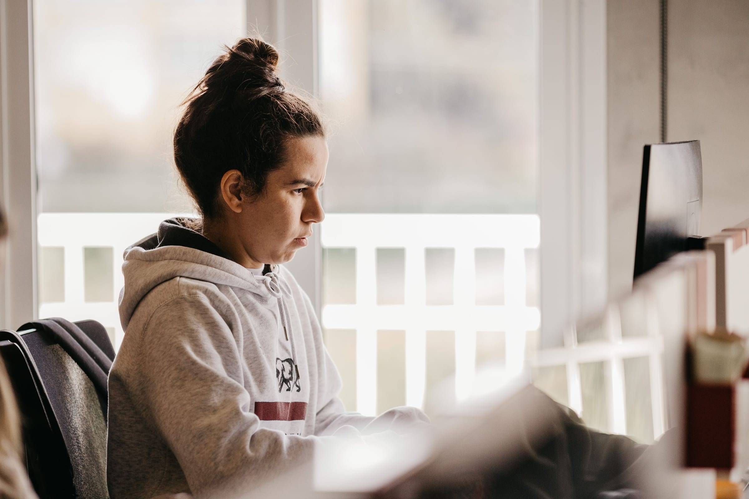 A person working at a screen by a window, a colleague passing out of focus in the foreground