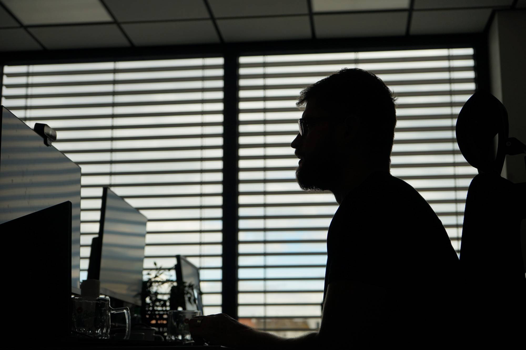 A Techmates developer silhouetted at their monitors against a window with blinds